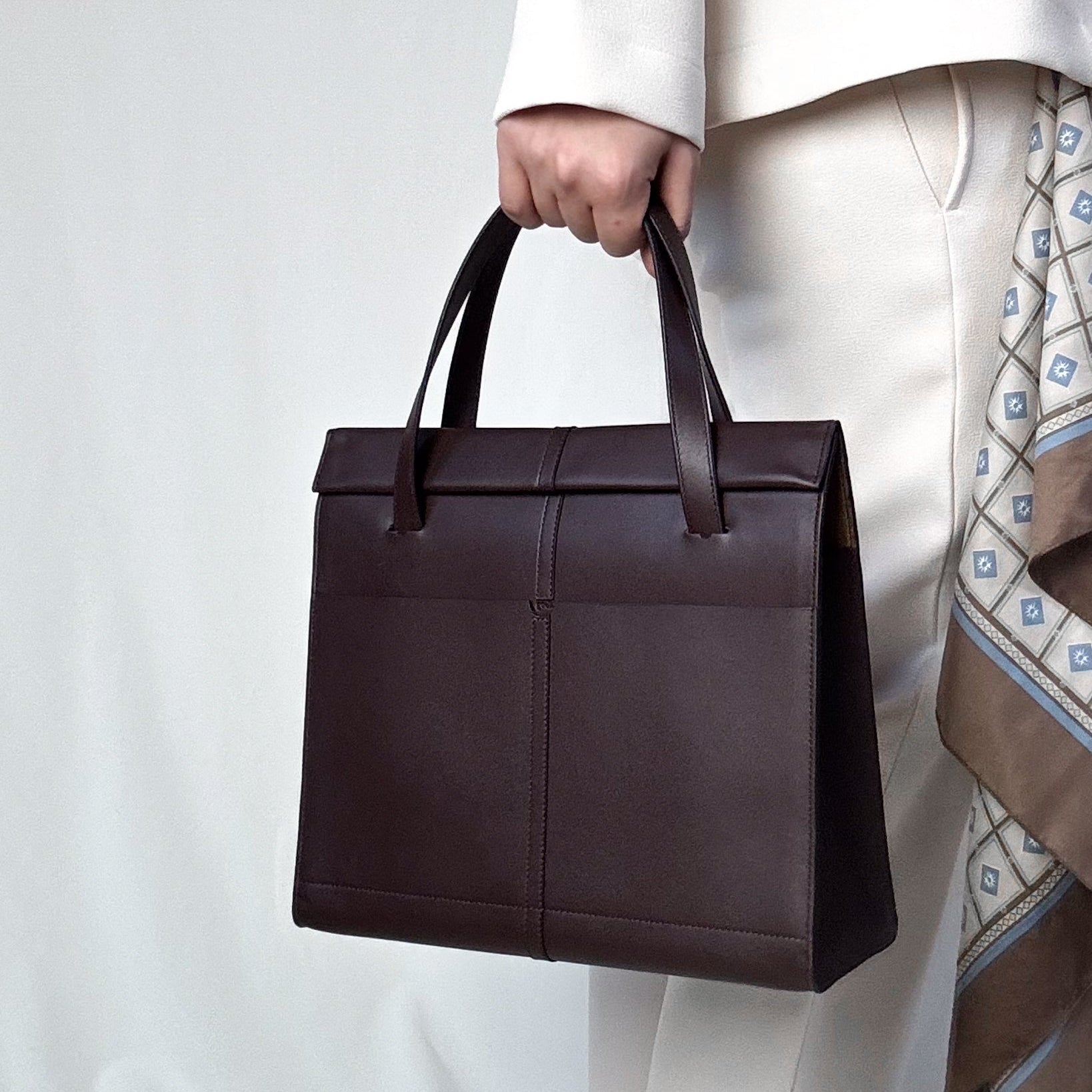 Brown leather bag held by a person wearing a white coat and patterned pants on a light gray background