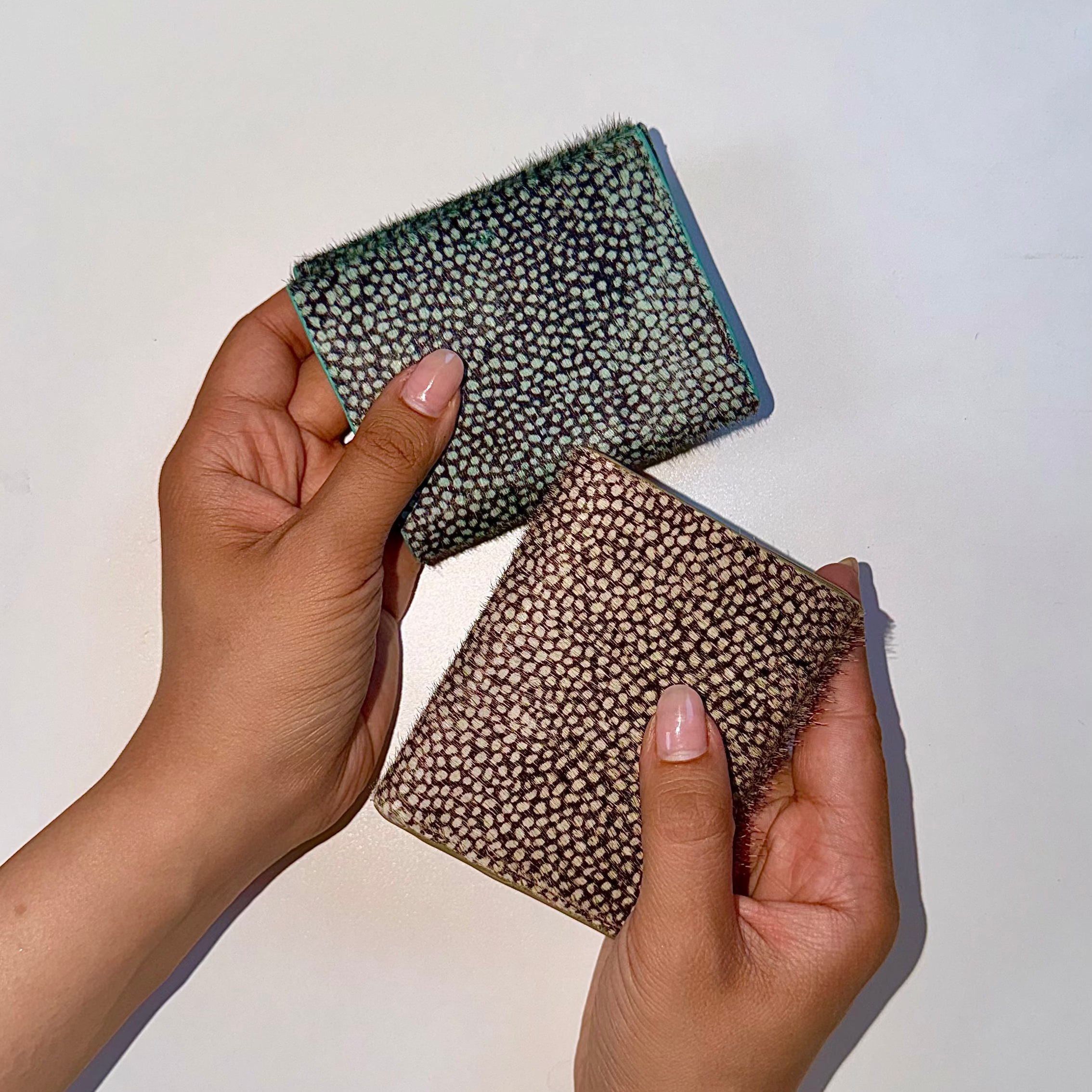 Two hands holding two small patterned wallets against a plain white background. One wallet has a dark green and black animal-print pattern with a blue edge, and the other has a brown and beige animal-print pattern.
